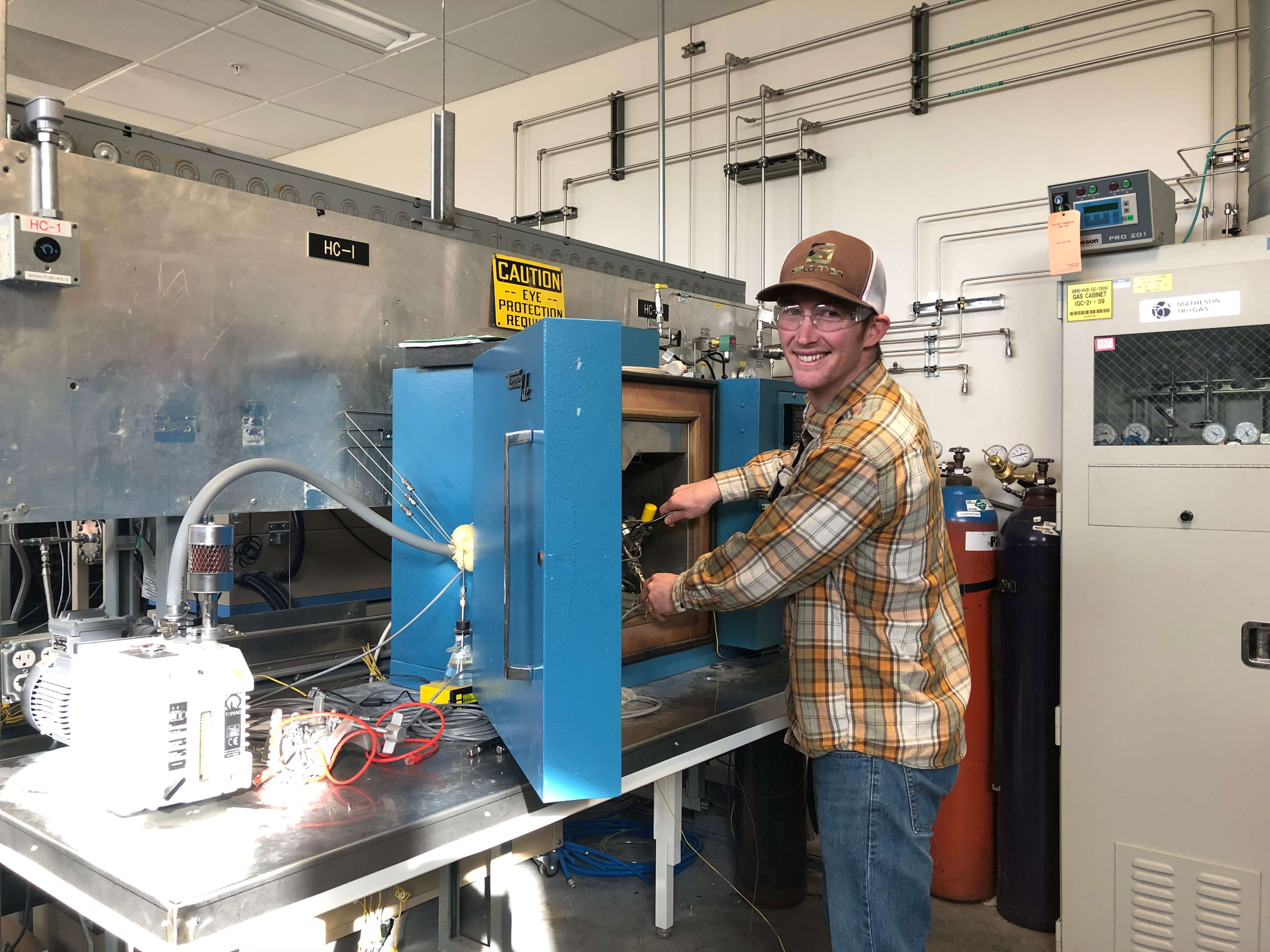 A smiling man uses a wrench to tighten something inside of a a large blue box. The boxes dore is open to allow access. Out of one side of the box, multiple tubes leave a sealed opening. One tube goes to a compressor, another goes to an erlenmeyer flask with a stopper, the rest go out of sight.